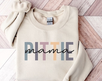 Puede incluir: Una sudadera de color crema con el texto "PIT TIE mama" en letras de bloque de colores. Las letras son en tonos de azul, morado, rosa, amarillo y verde.