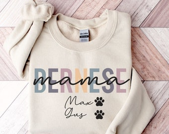Puede incluir: Sudadera color crema con la frase "BERNESE mama" en colores. Debajo, los nombres "Max" y "Jus" están impresos con dos huellas de patas. La sudadera tiene cuello redondo y mangas largas.