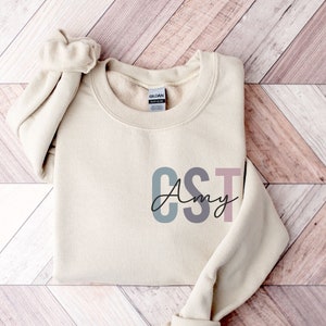 Könnte beinhalten: Ein beiger Pullover mit dem Text "CST Amy" in bunten Buchstaben. Die Buchstaben sind in Blautönen, Grau, Rosa und Braun.