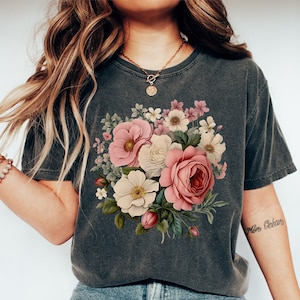 May include: A dark gray t-shirt with a floral print featuring pink, white, and green flowers. The flowers are arranged in a bouquet shape and are centered on the shirt. The shirt has a crew neck and short sleeves.