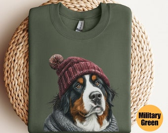 Puede incluir: Sudadera verde militar con un dibujo de un perro de montaña bernés con un gorro de punto burdeos y una bufanda gris. La sudadera se muestra sobre una superficie circular y tejida. El texto "Military Green" es visible.