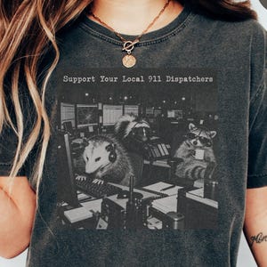 Puede incluir: Una camiseta gris carbón con un estampado gráfico en blanco y negro de una zarigüeya y un mapache con auriculares en un centro de emergencias 911. El texto sobre el gráfico dice Support Your Local 911 Dispatchers.