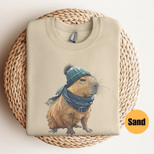 Puede incluir: Una sudadera beige con un gráfico de una capibara que lleva un gorro y una bufanda de punto azul y verde. La capibara está mirando hacia la derecha. La palabra "Sand" está impresa en negro sobre un círculo amarillo en la esquina inferior derecha de la imagen.