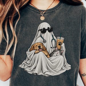 May include: A charcoal gray t-shirt featuring a graphic of a ghost wearing black sunglasses and a stethoscope around its neck. The ghost is holding an iced coffee and cuddling a golden retriever puppy and a small tabby cat under its white sheet.
