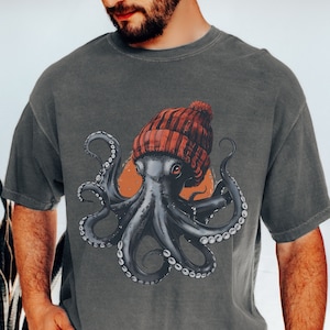 Op de afbeelding: Een donkergrijs T-shirt met een afbeelding van een octopus die een rood-witte muts draagt. De octopus heeft acht tentakels en is omringd door een lichtoranje cirkel.