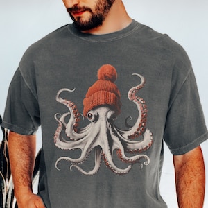 May include: Dark gray t-shirt featuring a detailed illustration of an octopus wearing a red knit beanie with a pom-pom. The octopus has white skin, red suckers, and large eyes. The shirt is a casual style.