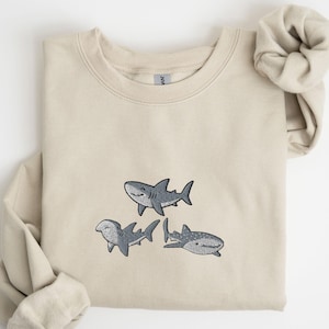 May include: A sand-colored sweatshirt featuring an embroidered design of three cartoon sharks in shades of gray. The word "Sand" is printed in black in the upper right corner. The sweatshirt has long sleeves and a crew neck.
