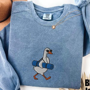 May include: Blue jean-colored sweatshirt with an embroidered goose wearing sunglasses and carrying a blue yoga mat. The sweatshirt has a crew neck and long sleeves. The text "Blue Jean" is in the bottom right corner.