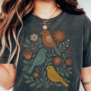 May include: Dark grey t-shirt featuring a colorful illustration of three birds perched among flowers and foliage. The birds are in shades of orange, blue, and yellow. The shirt has a vintage, worn look.