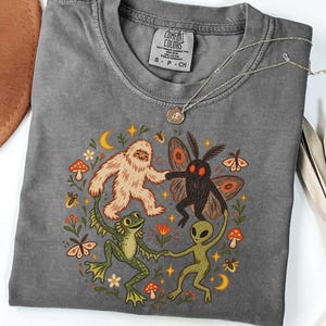 Può includere: Una t-shirt color pepe con una stampa grafica che raffigura uno yeti, una creatura falena, una rana e un alieno che si tengono per mano in cerchio. Il design include piccoli funghi, fiori, stelle e lune. Presenta un'etichetta Comfort Colors.
