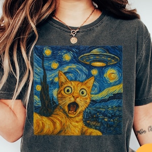 May include: Dark grey t-shirt featuring a vibrant, square graphic of a surprised orange tabby cat in front of a starry night sky with a UFO. The design is reminiscent of Van Gogh's style. The word "Pepper" is in a yellow circle.