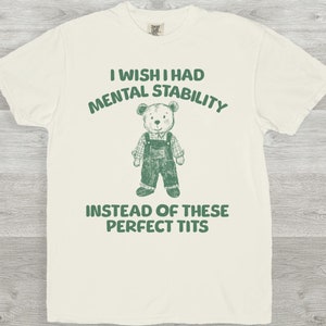 Könnte beinhalten: Elfenbeinfarbenes T-Shirt mit dem Text "I wish I had mental stability instead of these perfect tits." Ein Cartoon-Teddybär mit Latzhose befindet sich in der Mitte. Das Shirt besteht aus einem weichen Material.