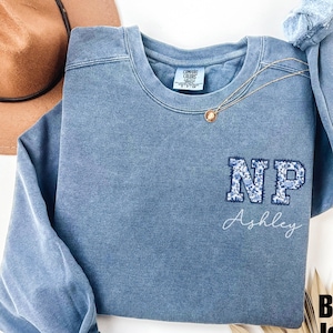 Könnte beinhalten: Blauer Jeans-Sweatshirt mit einem floralen "NP"-Monogramm und dem Namen "Ashley", der auf der Vorderseite gestickt ist.