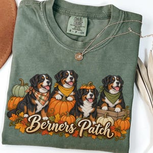 May include: A moss green t-shirt featuring a graphic of four Bernese Mountain Dogs sitting among pumpkins and autumn leaves. The dogs wear bandanas and one has a pumpkin on its head. The text "Berners Patch" is printed in a script font below.