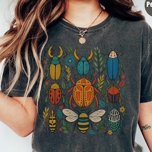 Pode incluir: Uma t-shirt cinzento-carvão com uma impressão gráfica colorida de vários escaravelhos e uma abelha rodeados por pequenas folhas e flores. O design inclui um grande escaravelho laranja central, uma abelha às riscas na parte inferior e vários outros insetos em tons de azul, amarelo e vermelho. A t-shirt tem gola redonda e mangas curtas.