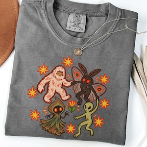May include: A pepper-colored t-shirt featuring a graphic print of a white yeti, a dark moth creature, a green alien, and a hooded figure holding hands. The design includes orange star accents and a small flower. The shirt has a Comfort Colors label.