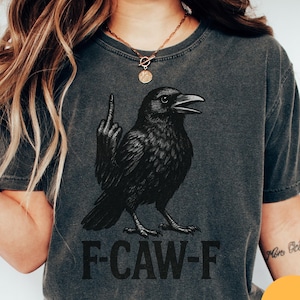 Puede incluir: Camiseta gris oscuro con una ilustración de un cuervo negro haciendo una peineta. El texto "F-CAW-F" está impreso debajo del cuervo. La camiseta tiene un aspecto vintage y desgastado.