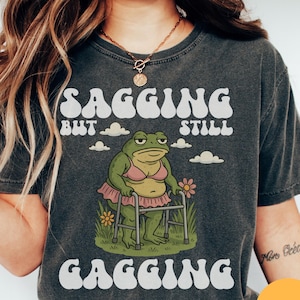 May include: Dark grey t-shirt with the text "Sagging But Still Gagging" in white. The shirt features a cartoon frog wearing a pink bra and a pink tutu, using a walker. The frog is in a grassy area with flowers.