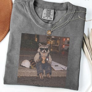 Puede incluir: Una camiseta de cuello redondo color pimienta con un estampado gráfico de un mapache con gafas de sol y peto, sosteniendo una bebida. El mapache está sentado en un bordillo junto a un casco blanco, una cinta métrica y un portapapeles.