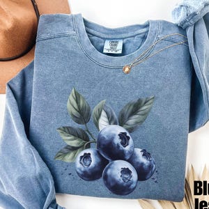 Op de afbeelding: Een blauw jeans sweatshirt met een afbeelding van blauwe bessen aan een tak. Het sweatshirt heeft een Comfort Colors label aan de voorkant.