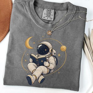Puede incluir: Una camiseta gris carbón con un gráfico de un astronauta en traje blanco sentado leyendo un libro. El diseño incluye una luna creciente amarilla, un planeta pequeño y estrellas. La camiseta tiene una etiqueta Comfort Colors en el cuello.