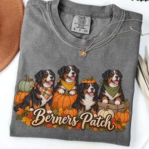 以下が含まれることがあります： ペッパーカラーのTシャツで、カボチャや秋の葉の中に座っている4匹のバーニーズ・マウンテンドッグのグラフィックが描かれています。犬たちはカラフルなバンダナを巻いており、1匹はカボチャの帽子をかぶっています。Berners Patchという文字が筆記体でプリントされています。