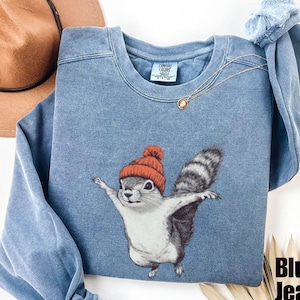 May include: A blue jean sweatshirt with a graphic of a squirrel wearing a red winter hat. The squirrel is flying through the air with its arms outstretched.