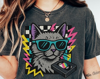 Op de afbeelding: Donkergrijs t-shirt met een retro afbeelding van een kat met een zonnebril en een VHS-band. Het ontwerp bevat een geblokte achtergrond, bliksemflitsen en het woord "Pepper" in een gele cirkel.