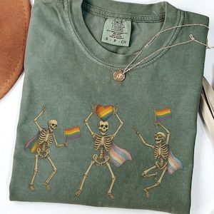 May include: Moss green t-shirt with a graphic of three dancing skeletons wearing rainbow capes and holding rainbow flags and a rainbow heart. A silver necklace with a pendant is on the shirt. The shirt has a Comfort Colors tag.