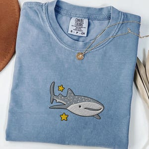 May include: A blue jean coloured t-shirt with an embroidered whale shark design and two yellow stars. The shirt has a crew neck and short sleeves. A gold necklace with a pendant is draped across the shirt.