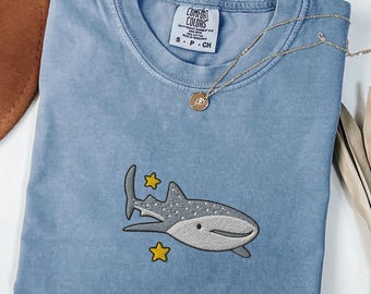 Op de afbeelding: Een blauw spijkerbroekkleurig t-shirt met een geborduurd walvishaai-ontwerp en twee gele sterren. Het shirt heeft een ronde hals en korte mouwen. Een gouden ketting met een hanger is over het shirt gedrapeerd.