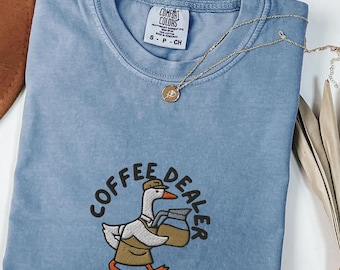 May include: A blue jean colored t-shirt with a crew neckline and a Comfort Colors label. The shirt features an embroidered graphic of a goose wearing a brown apron and cap, holding a coffee pot, with the text COFFEE DEALER arched above.