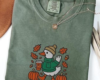 May include: A moss green Comfort Colors t-shirt featuring an embroidered duck wearing a hat, scarf, and boots, holding a coffee cup, surrounded by pumpkins and fall leaves. A gold necklace with a pendant is also visible.
