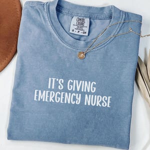 May include: A blue jean Comfort Colors t-shirt with the text "IT'S GIVING EMERGENCY NURSE" in white. A gold necklace with a pendant is draped across the shirt. The shirt is folded and ready to wear.