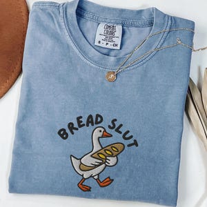 May include: Blue jean Comfort Colors t-shirt with the embroidered phrase "BREAD SLUT" and a cartoon duck holding a loaf of bread. A gold necklace with a pendant rests on the shirt. The shirt is size S-P-CH.