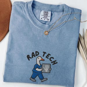 May include: Blue jean colored t-shirt with the text "RAD TECH" and a cartoon duck wearing scrubs and holding an x-ray. The shirt has a crew neck and short sleeves. A gold necklace with a pendant is draped across the shirt.