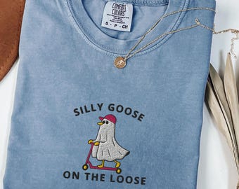 Op de afbeelding: Blauw spijkerbroekkleurig T-shirt met de tekst "Silly Goose on the Loose" en een afbeelding van een gans op een scooter. Het shirt heeft een ronde hals en korte mouwen. Een gouden ketting met een hanger ligt over het shirt.