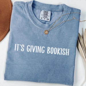 May include: A blue jean-colored t-shirt with the phrase "IT'S GIVING BOOKISH" in white block letters. A gold necklace with a small pendant rests on the shirt. The shirt is folded, and the tag reads "Comfort Colors".