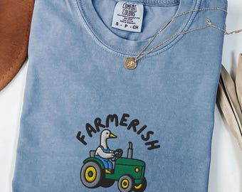 May include: Blue jean Comfort Colors t-shirt with a crew neck. The shirt features a graphic of a duck driving a green tractor with the word "Farmerish" above it. A gold necklace with a pendant is draped across the shirt.