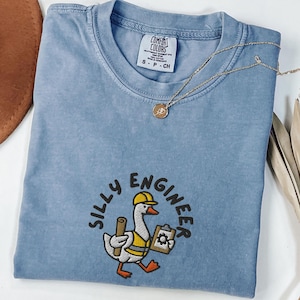 Peut inclure: Un t-shirt bleu jean Comfort Colors avec le texte "SILLY ENGINEER" et un dessin d'oie portant un casque et des plans. Un collier en or avec un pendentif est posé sur le t-shirt.