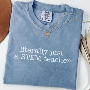 May include: A blue jean colored t-shirt with the text "literally just a STEM teacher" in white. A gold necklace with a pendant is draped across the shirt. The shirt is folded and has a Comfort Colors tag.