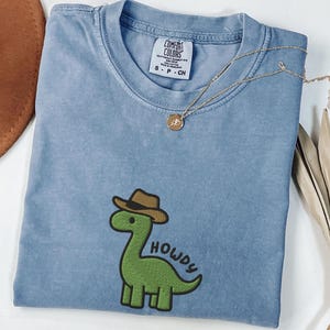 Puede incluir: Una camiseta azul Comfort Colors con un dinosaurio verde con un sombrero de vaquero marrón. La palabra "HOWDY" está escrita en blanco. Un collar dorado con un colgante está sobre la camisa.