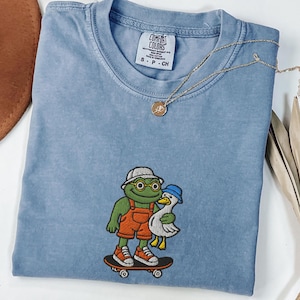 May include: Blue jean Comfort Colors t-shirt with an embroidered design of a frog and a duck on a skateboard. The frog wears glasses, a hat, and orange overalls. A gold necklace with a pendant rests on the shirt.