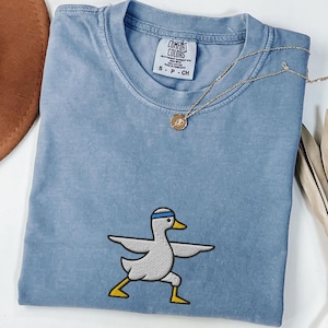 Könnte beinhalten: Ein blaues Jeans-T-Shirt mit einer Cartoon-Ente, die Yoga macht. Das Shirt hat einen Rundhalsausschnitt und kurze Ärmel. Eine goldene Halskette mit einem kleinen Anhänger ist über dem Shirt drapiert. Das Shirt hat das Comfort Colors-Label.