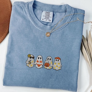 Op de afbeelding: Blauw spijkerbroekkleurig T-shirt met een geborduurd ontwerp van vier geesten, elk met unieke accessoires. Het shirt heeft een ronde hals en korte mouwen. Een gouden ketting met een hanger ligt op het shirt. De tekst "Blue Jean" is zichtbaar.