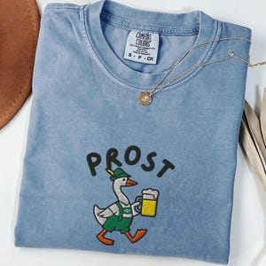 May include: A blue jean Comfort Colors t-shirt with the word "PROST" above an embroidered goose wearing a hat and lederhosen, holding a beer stein. A gold necklace with a pendant is draped across the shirt.