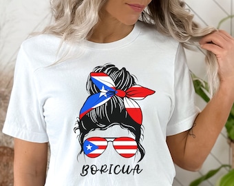 Puede incluir: Camiseta blanca con un gráfico de una mujer con un moño desordenado, usando gafas de sol con la bandera puertorriqueña en ellas. El moño está decorado con una cinta roja, blanca y azul con la bandera puertorriqueña en ella. El texto "BORICUA" está impreso debajo del gráfico.