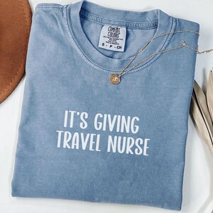 Peut inclure: Un t-shirt bleu jean Comfort Colors avec le texte "IT'S GIVING TRAVEL NURSE" en blanc. Un collier en or avec un pendentif est drapé sur le t-shirt. Le t-shirt est plié et posé sur une surface blanche.