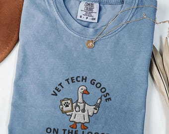 Puede incluir: Camiseta azul vaquero Comfort Colors con el texto "VET TECH GOOSE ON THE LOOSE" y un dibujo de un ganso con bata de laboratorio, sujetando una carpeta y una jeringuilla. Un collar dorado con un colgante está sobre la camiseta.
