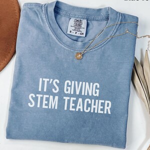 May include: A blue jean colored t-shirt with the text "IT'S GIVING STEM TEACHER" in white. A gold necklace with a pendant is draped across the shirt. The shirt is folded and has a Comfort Colors tag.